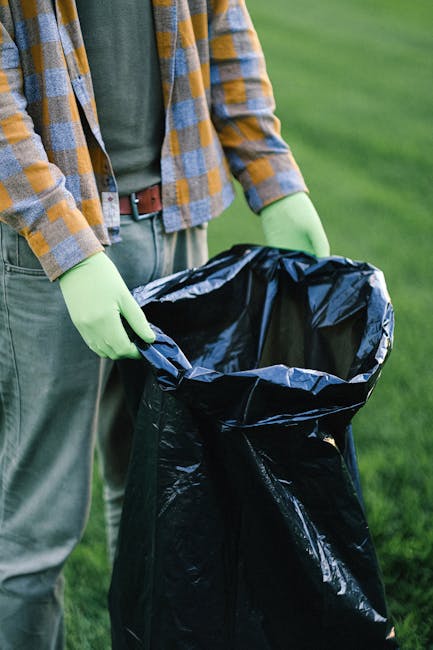 A sanitation worker wearing a high-visibility orange vest and black trousers is operating a large red garbage truck parked on the side of a road. The worker is seen leaning slightly forward, using the