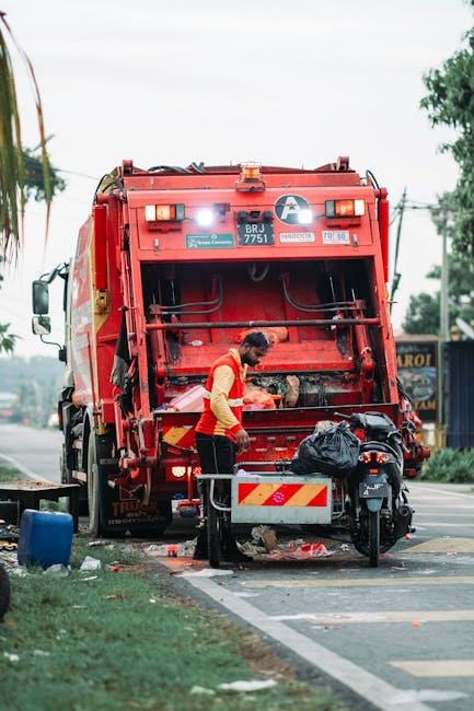 A waste collection operative in a bright orange high-visibility vest and dark trousers is emptying a black plastic bag into the rear compactor of a large, red waste collection truck parked on the side
